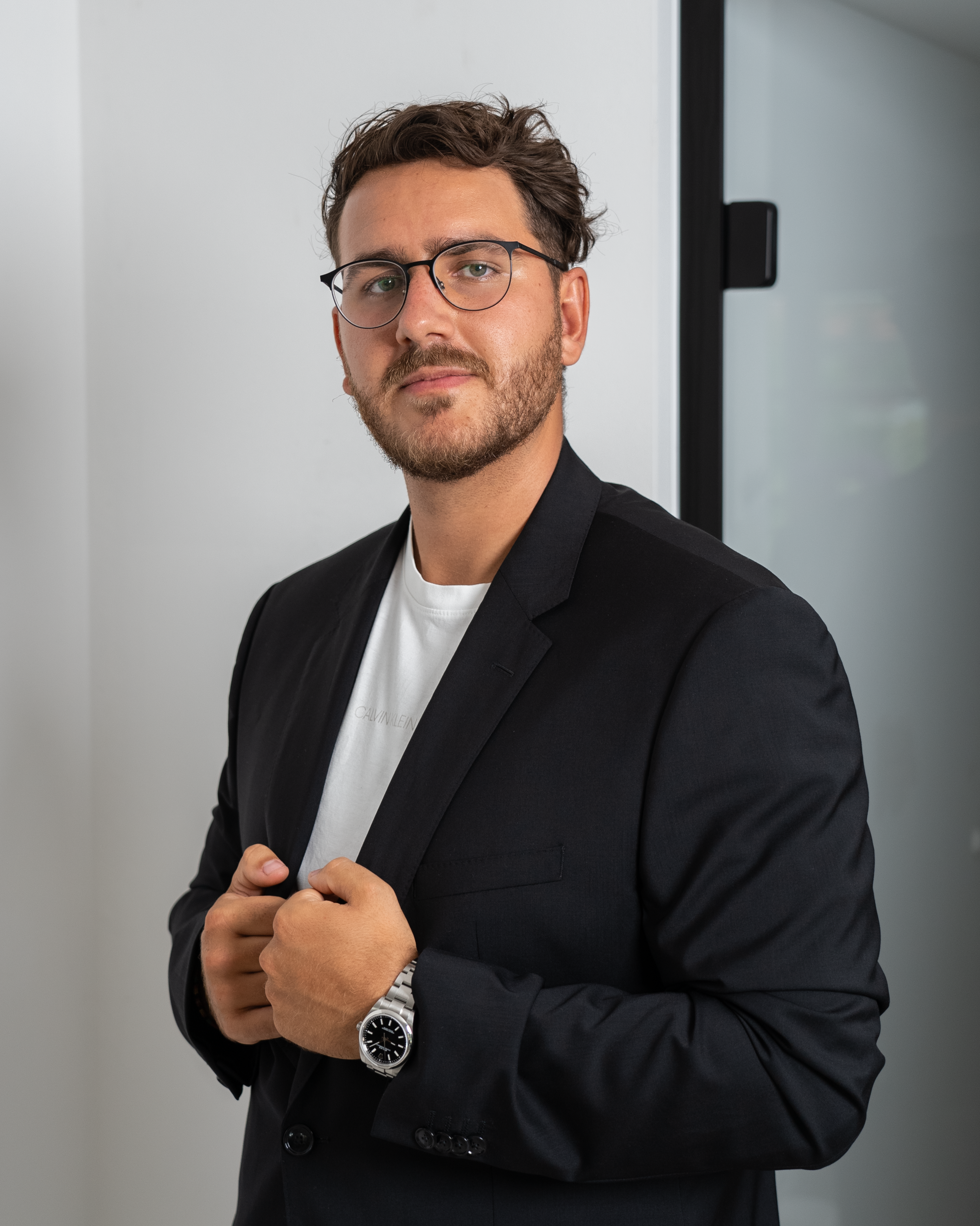 Portrait of a confident man in a black suit, wearing glasses and standing indoors