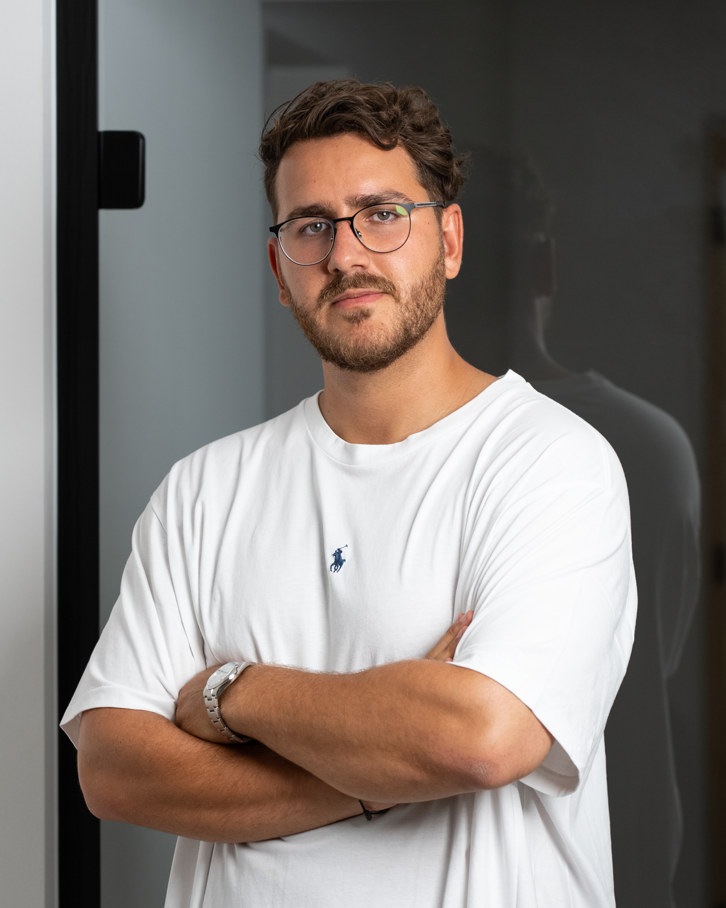 Portrait of a confident man in a white t-shirt and eyeglasses with arms crossed indoors.
