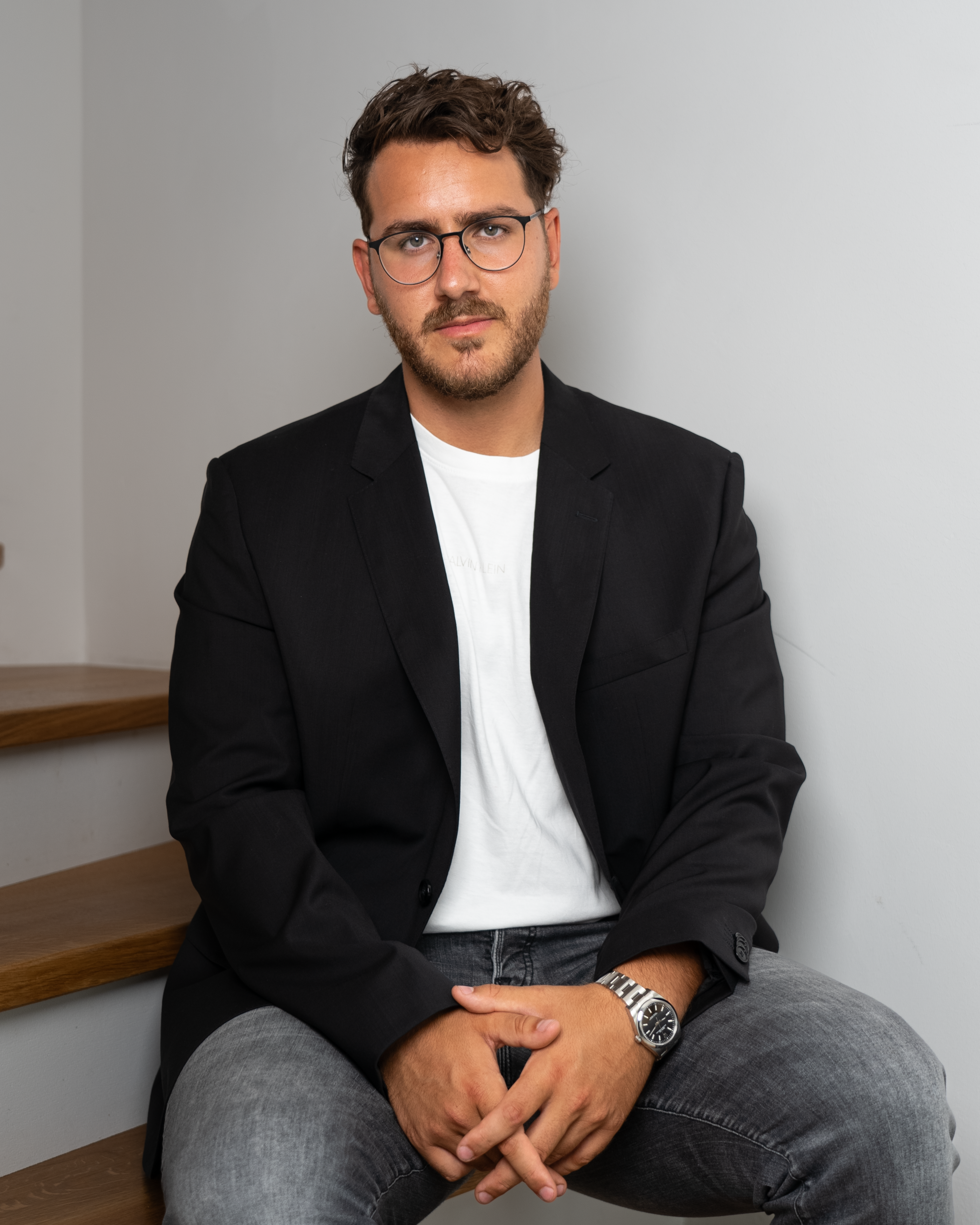 Elegant and modern portrait of a man in a black suit jacket and eyeglasses, sitting indoors.