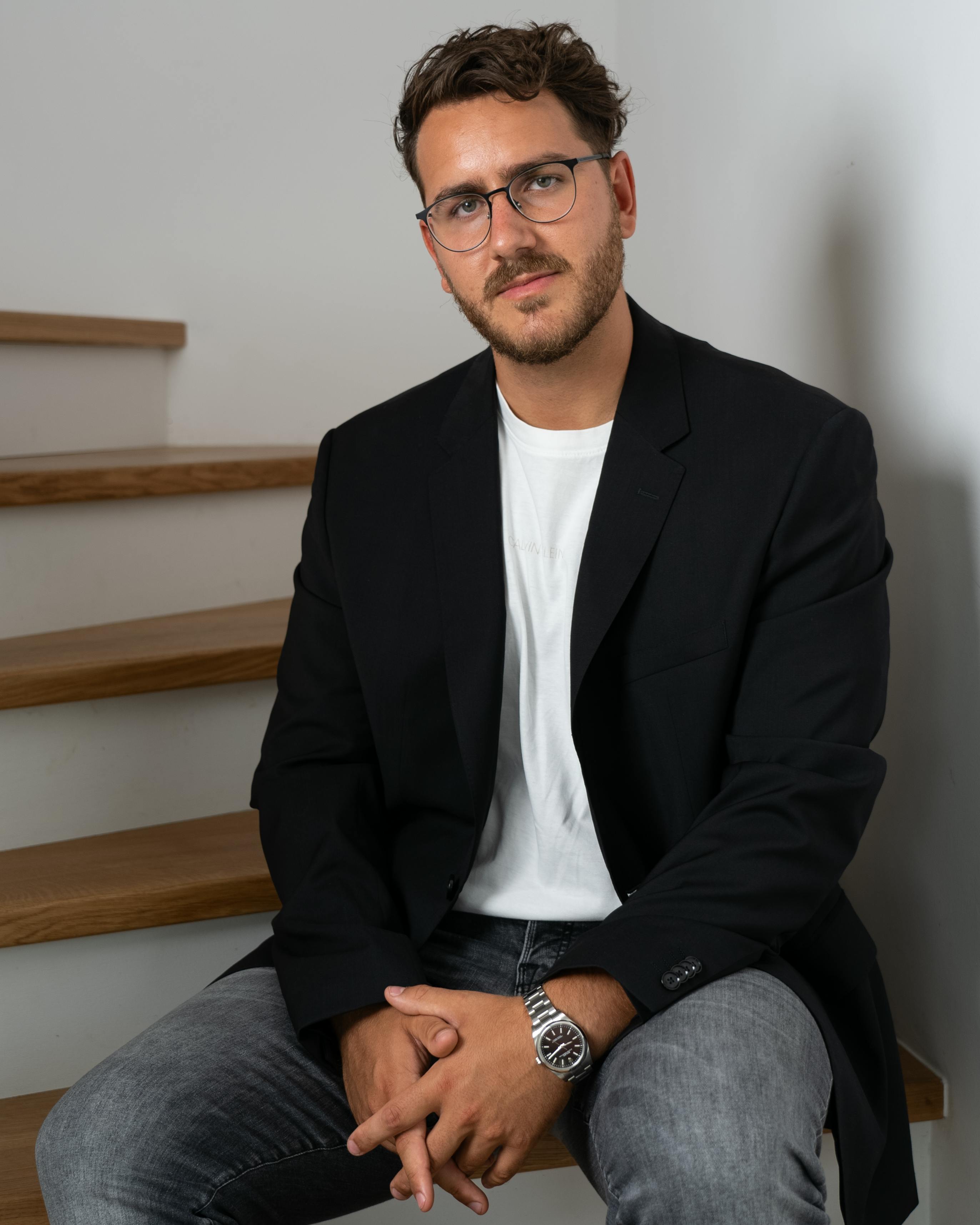 A stylish adult man in casual business attire sitting on stairs indoors.