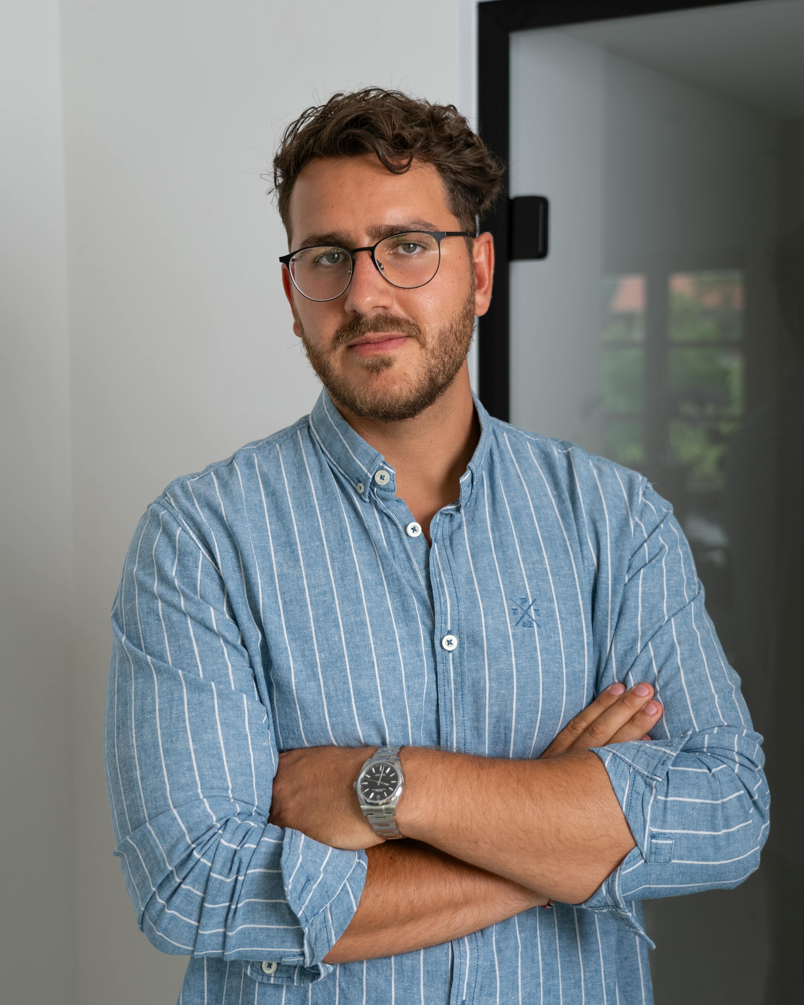 Well-dressed man with eyeglasses and stubble stands confidently indoors, arms crossed.