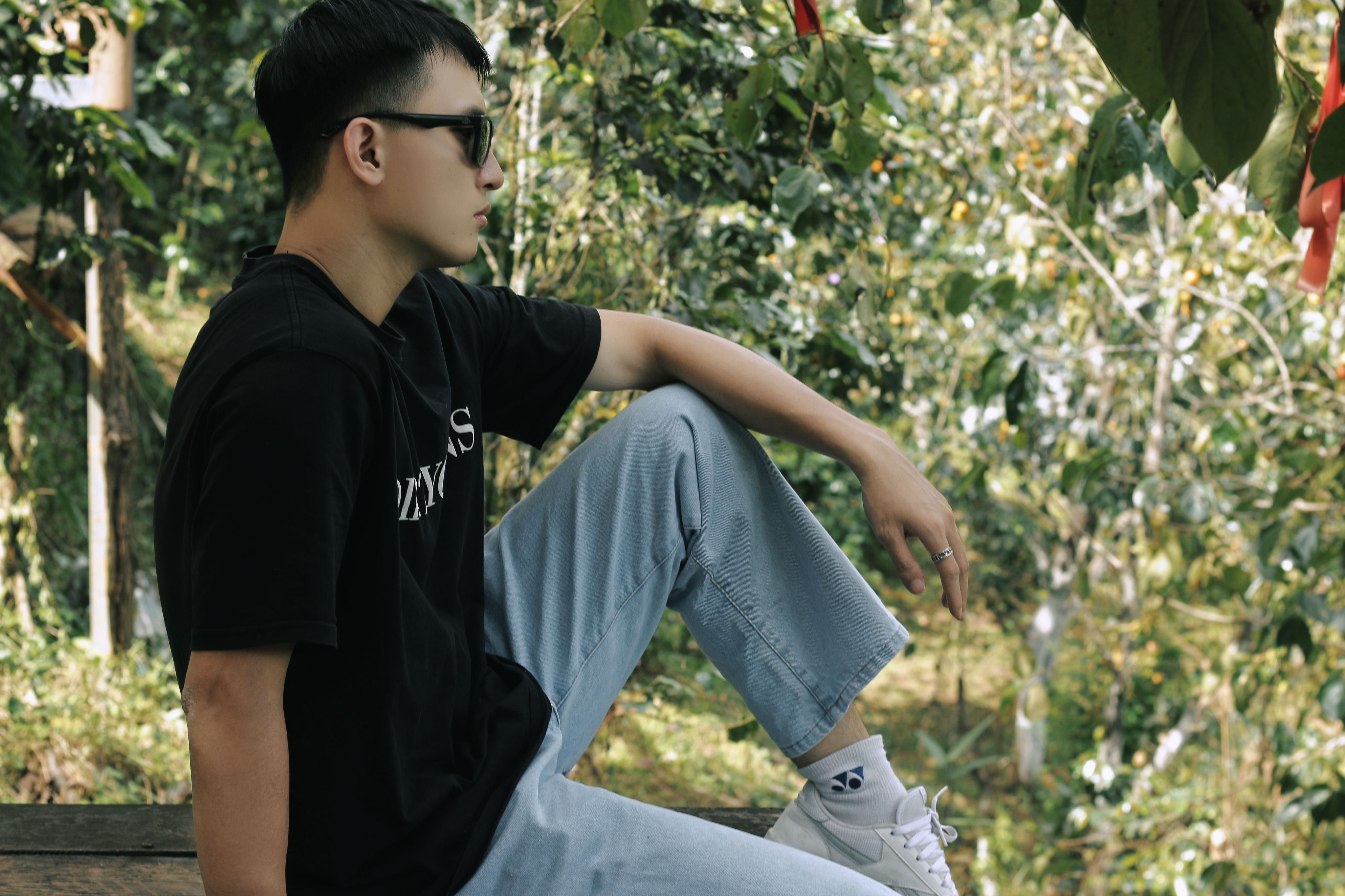 Stylish man in sunglasses and black t-shirt sitting outdoors surrounded by greenery.