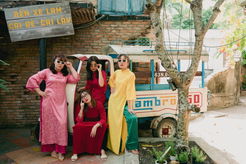 Group of young women wearing ao dai and sunglasses posing enthusiastically by an old vehicle in Vietnam.