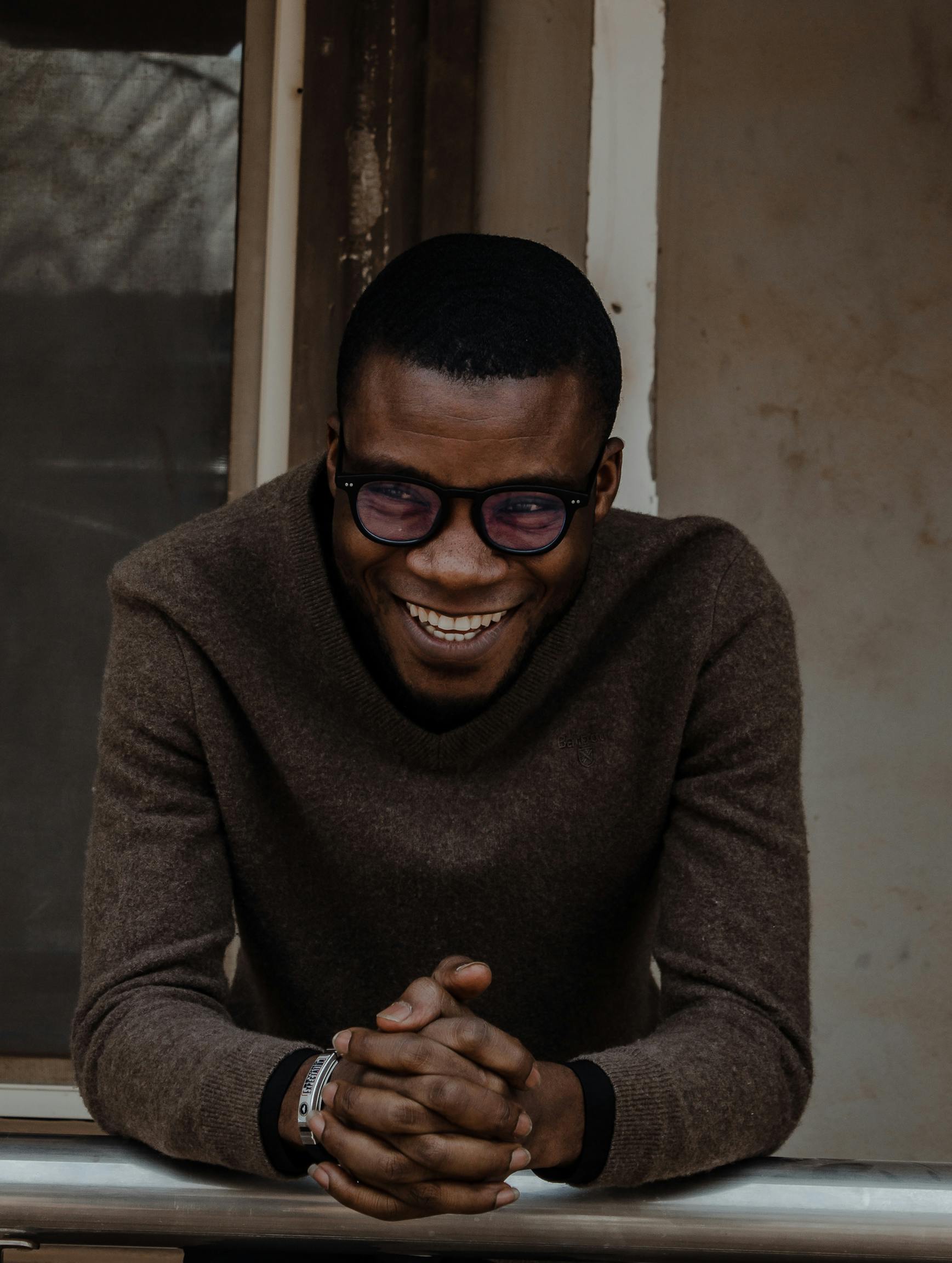 A cheerful young man wearing a brown woolen sweater and eyeglasses, leaning and smiling warmly.