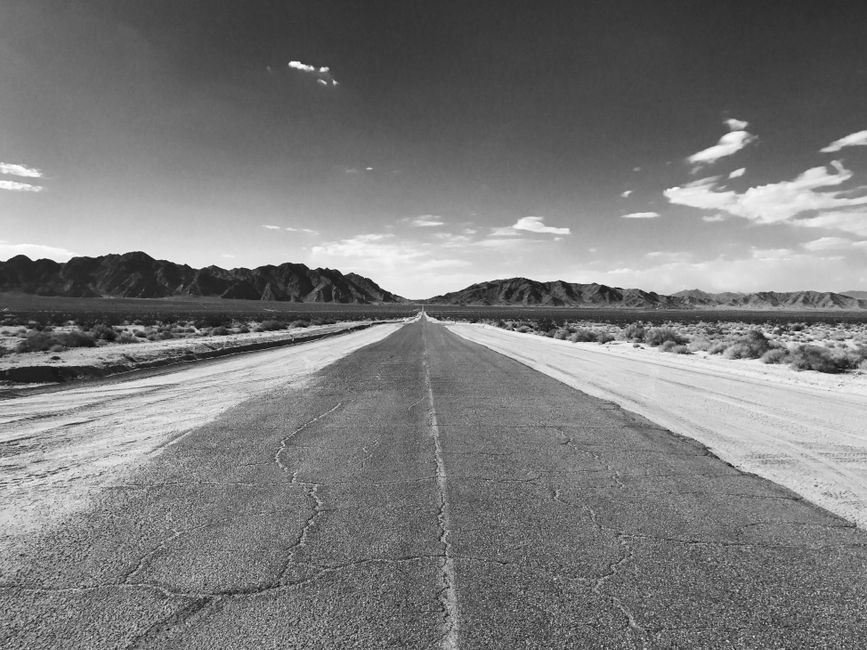 A dramatic black and white view of an empty highway stretching through a desert landscape under a cloudy sky.