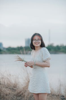 Young Asian woman with glasses smiling by a lake, holding grass on a cloudy day.