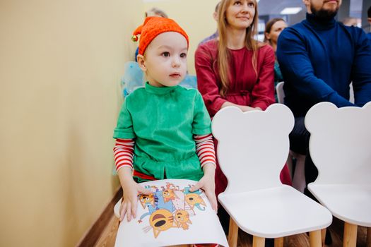 A young child dressed as an elf sits at a Christmas event indoors.