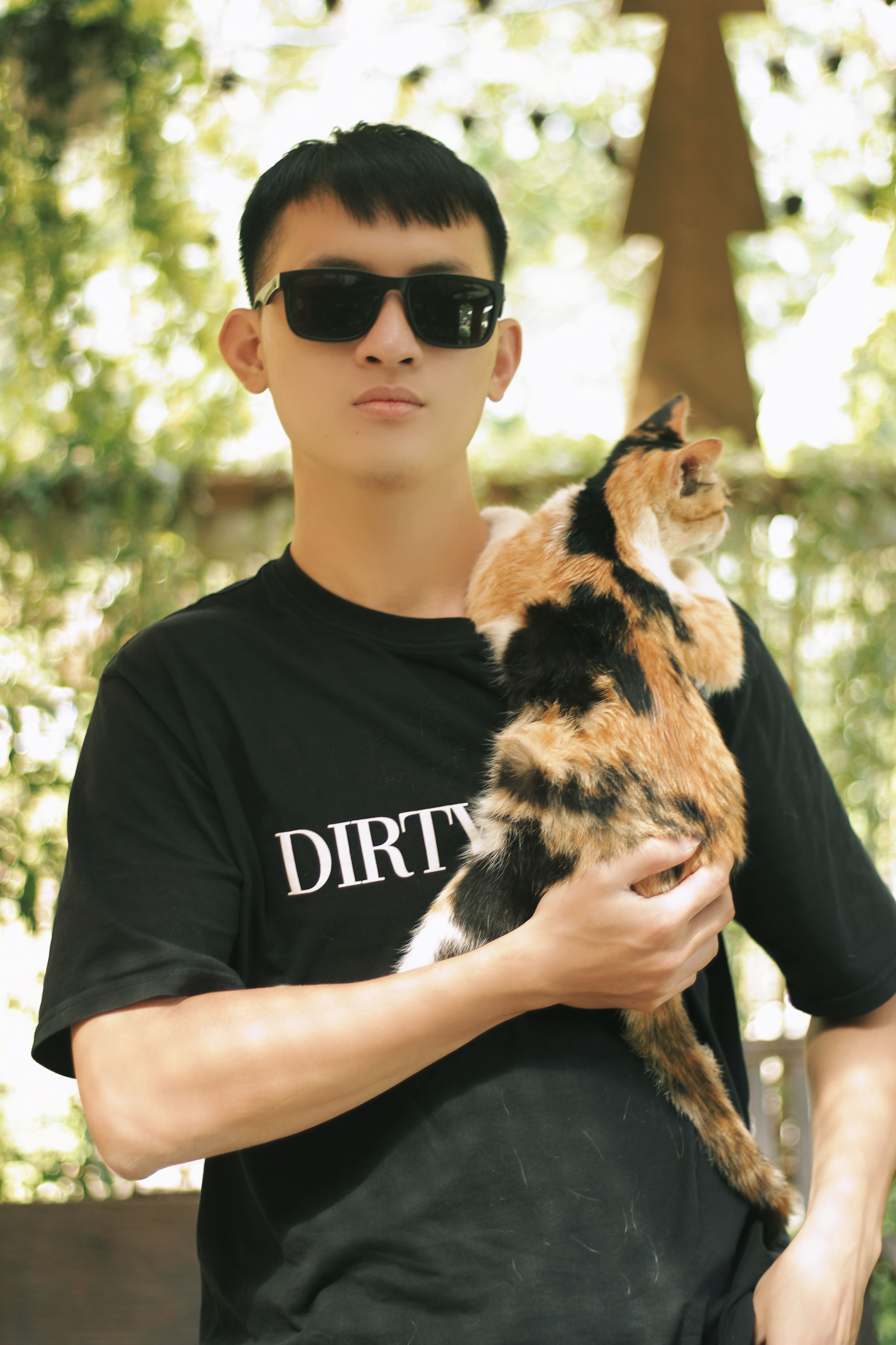 A fashionable young man holding a calico cat outdoors on a sunny day.