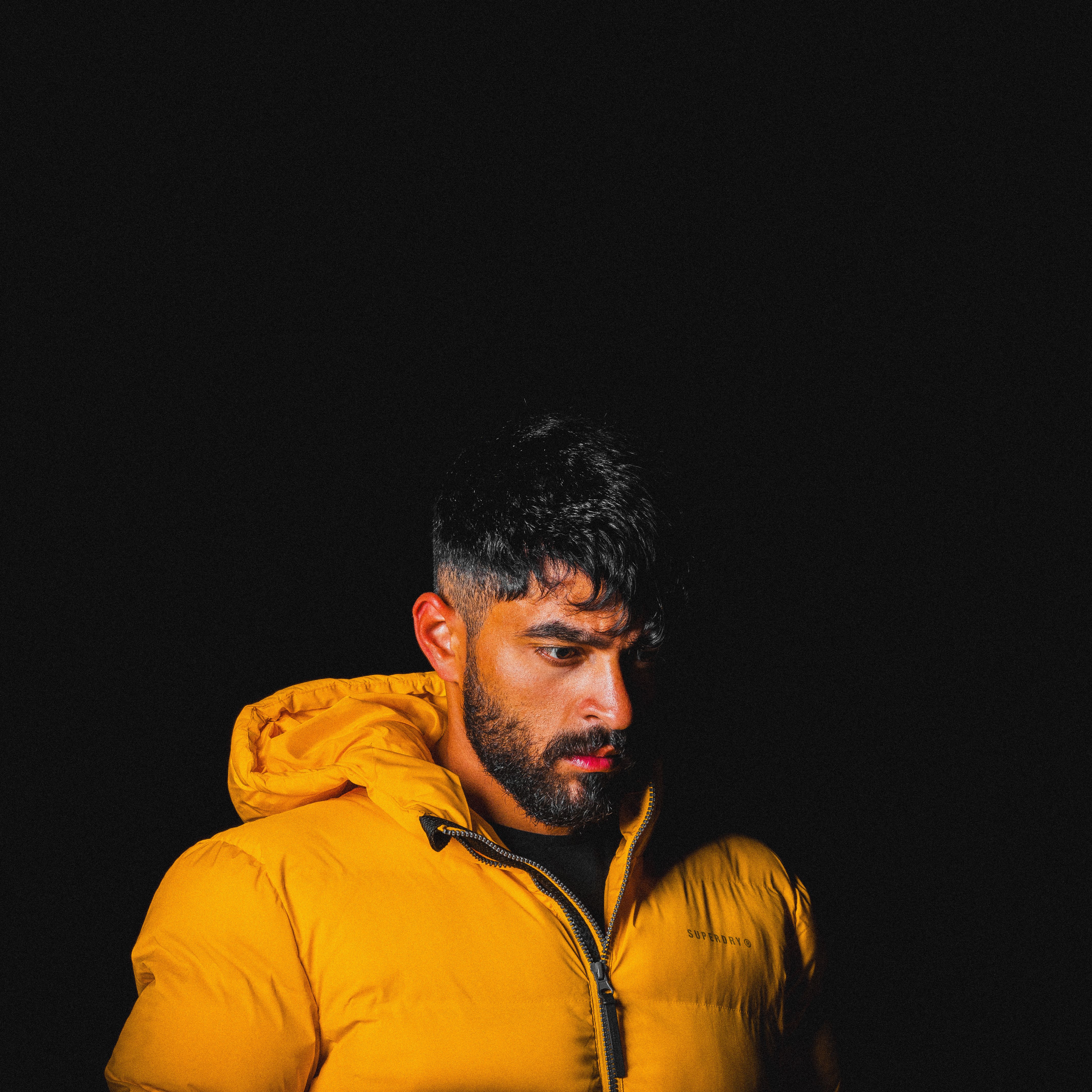 Dramatic portrait of a man with beard wearing a yellow jacket against a dark background.