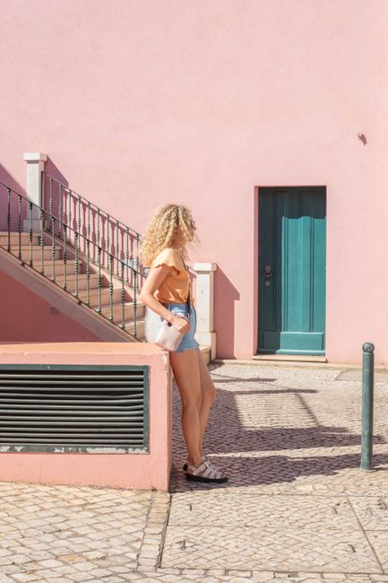 Stylish woman in urban setting beside pink building with turquoise door on a sunny day.
