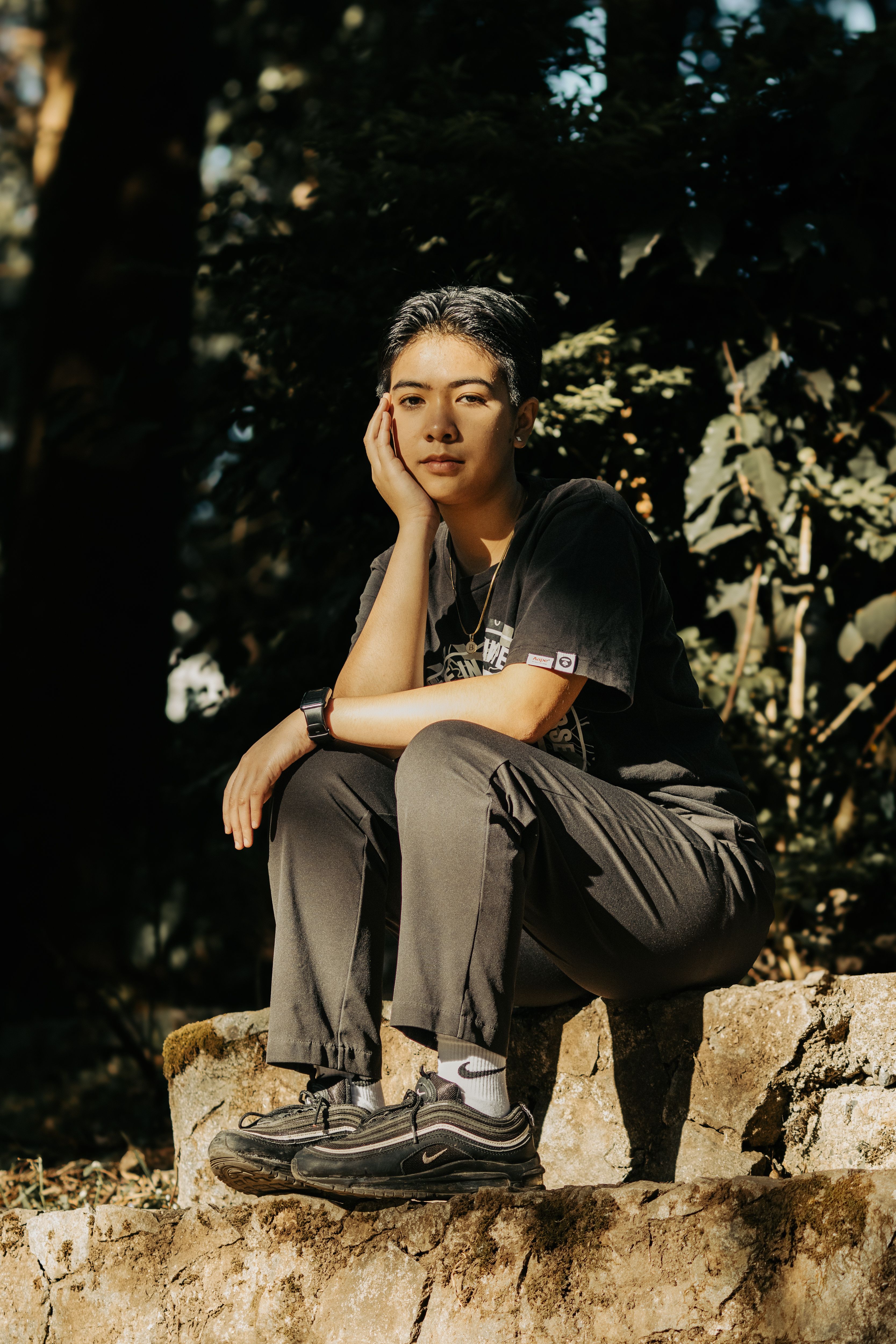 Stylish young woman sitting outdoors, wearing casual attire, enjoying a sunny day in nature.