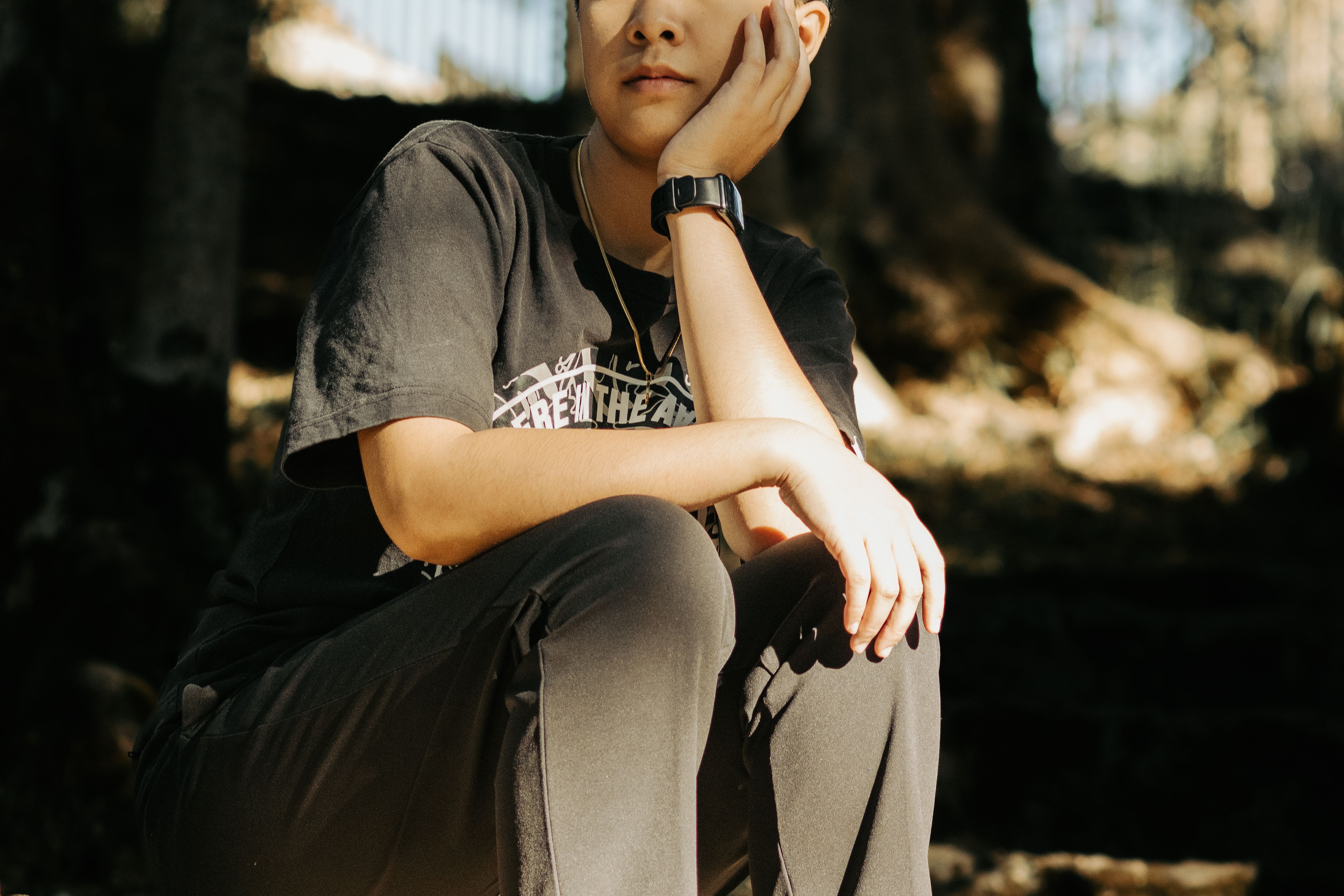 A person in a black T-shirt sits outdoors with a thoughtful expression, hand on cheek.