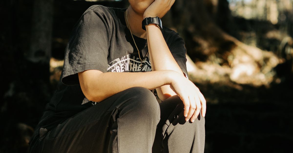 A person in a black T-shirt sits outdoors with a thoughtful expression, hand on cheek.