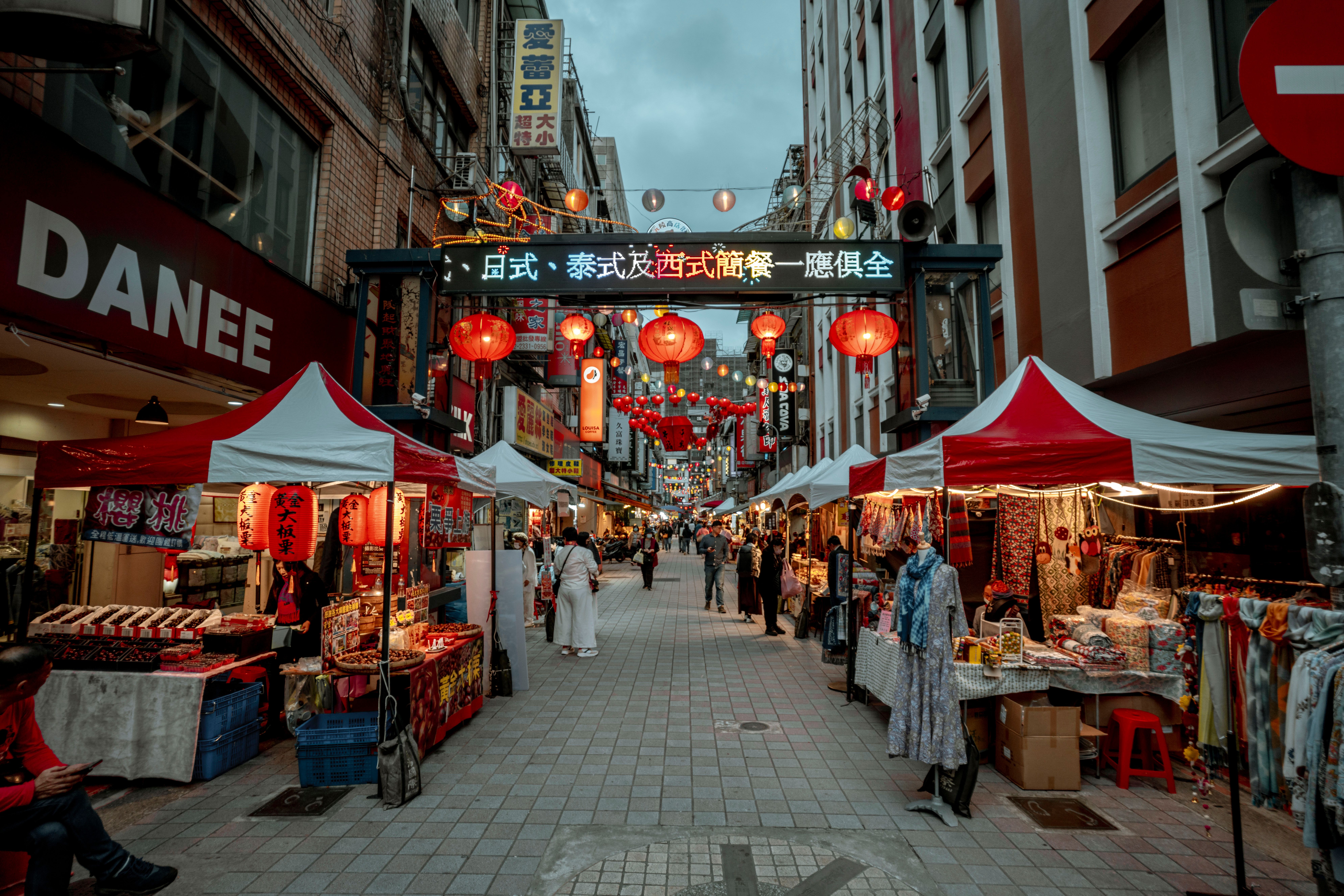 Bustling Asian street market with red lanterns and vibrant stalls offering goods.