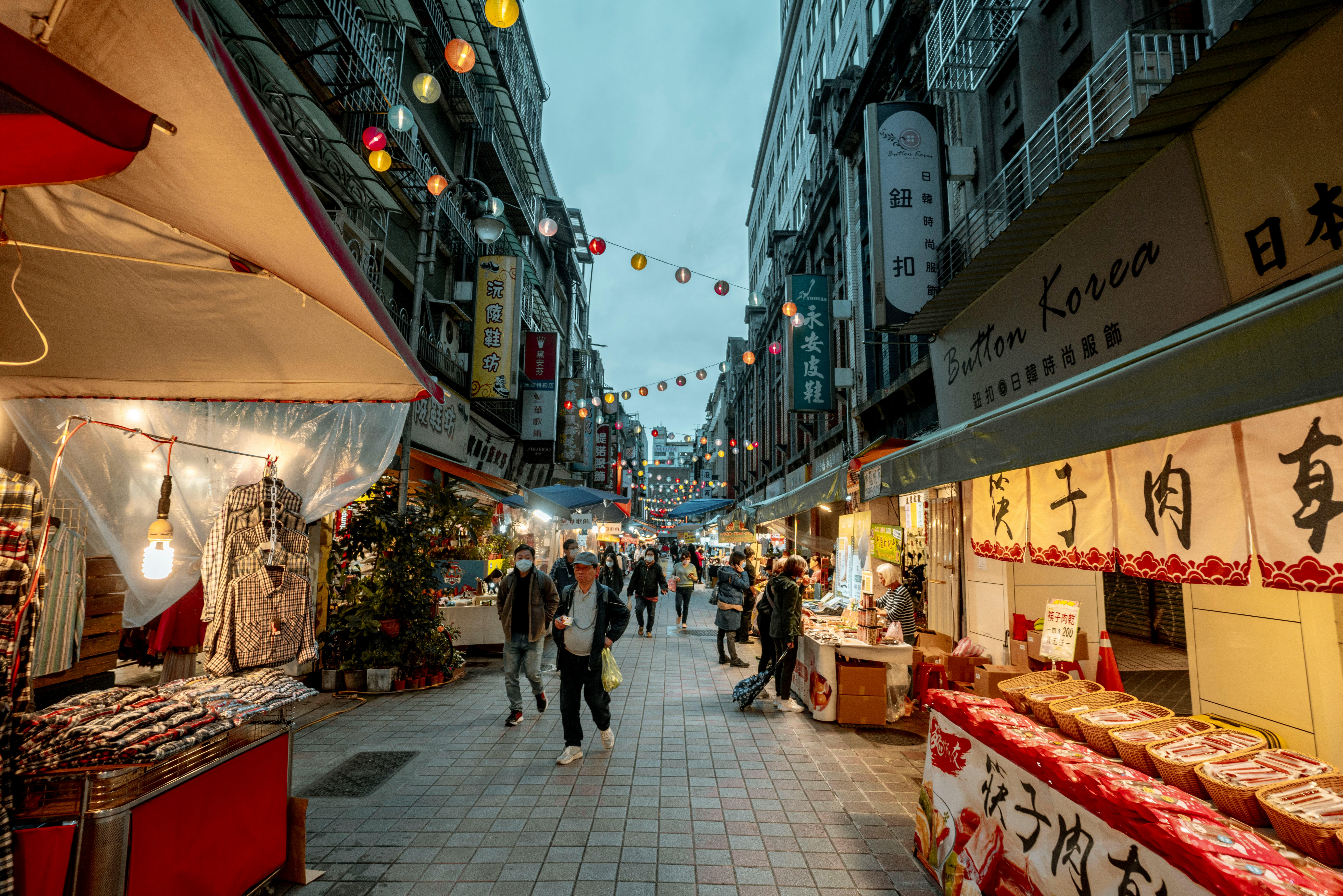 Busy street market in Asia at dusk, featuring shops, lanterns, and people shopping.