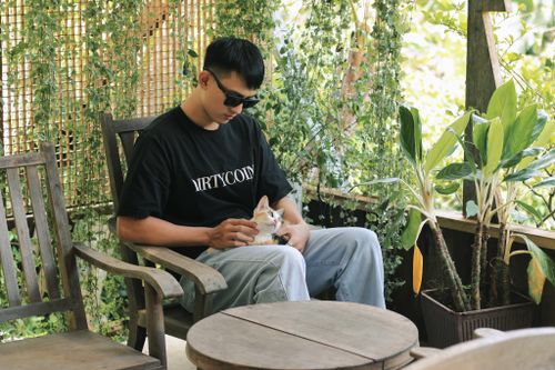 Free Asian man in sunglasses gently pats a cat while sitting outdoors in a lush green garden setting. Stock Photo