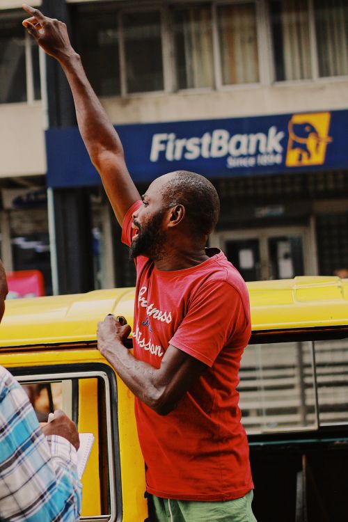 Free A man in a red t-shirt raises his arm in an urban street setting. FirstBank sign visible. Stock Photo