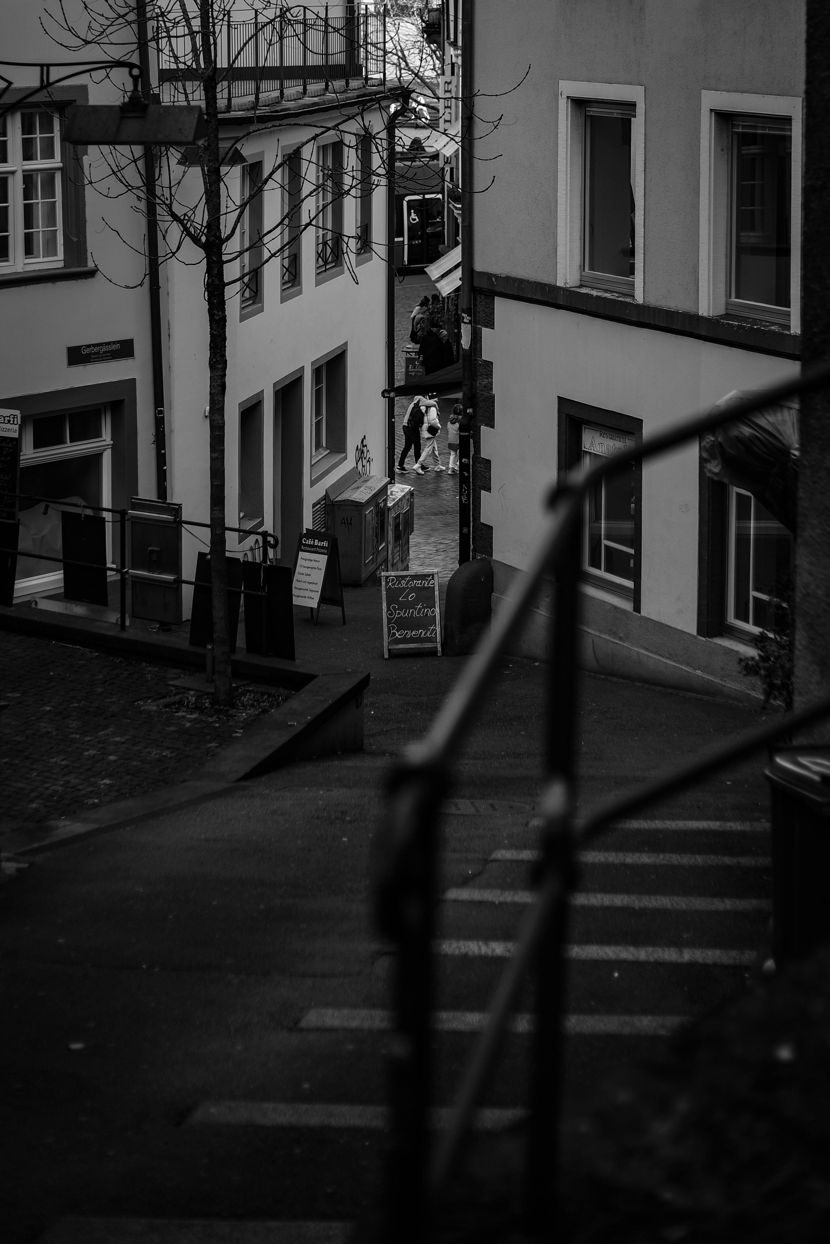 A moody black and white view of an urban alleyway with buildings and people in the distance.