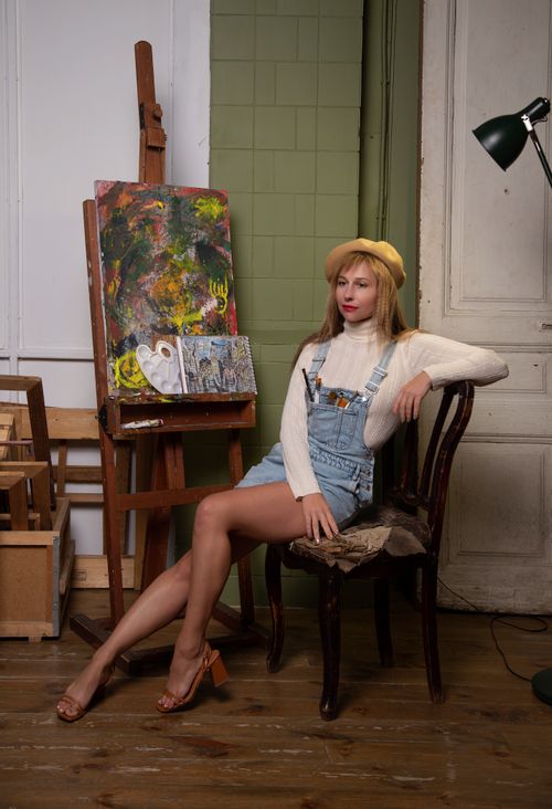 Free A young woman artist in denim overalls poses beside an easel in a vintage art studio. Stock Photo