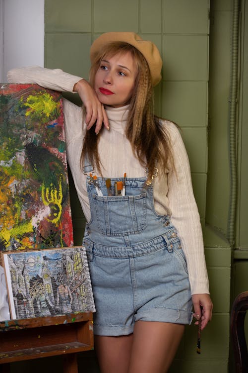 A woman in overalls and a hat posing next to a painting