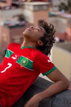 A young man in a red Moroccan soccer jersey leans back with eyes closed in an urban setting.