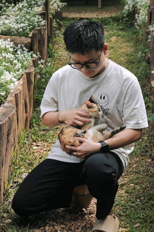 Free A young man cuddles a cat while kneeling in a picturesque garden pathway during summer. Stock Photo