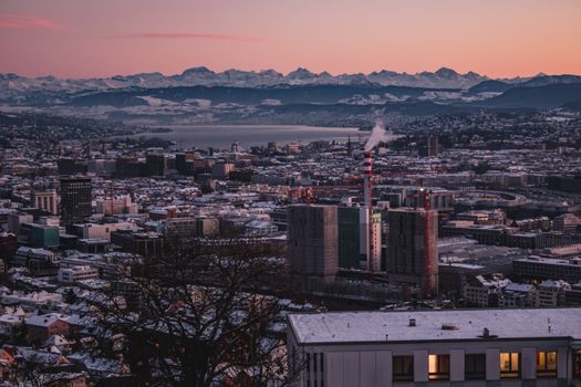 Aerial view of Zürich cityscape at winter sunset with snowy Alps backdrop.