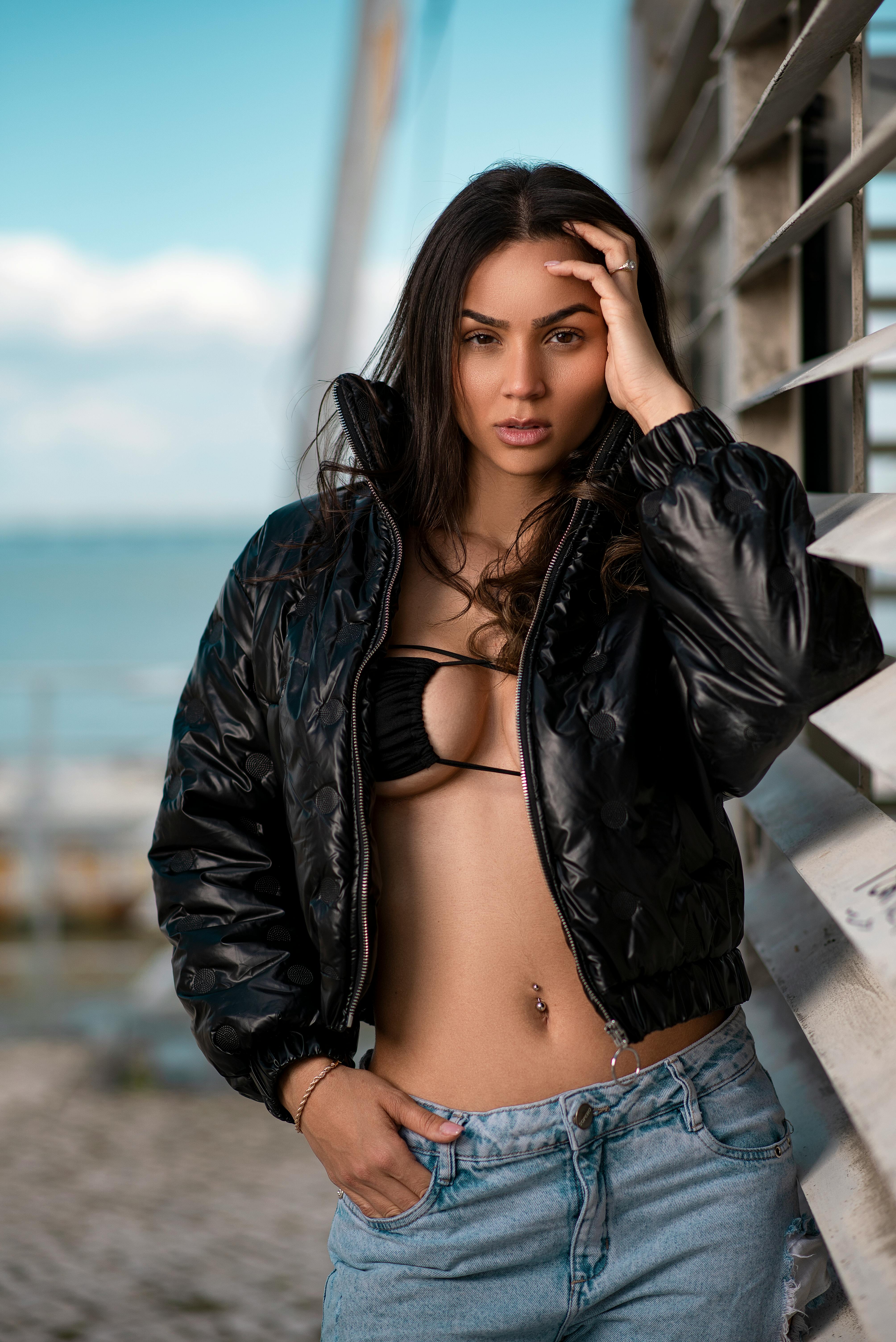 Fashionable woman in a black jacket poses by a railing with the sea in the background.