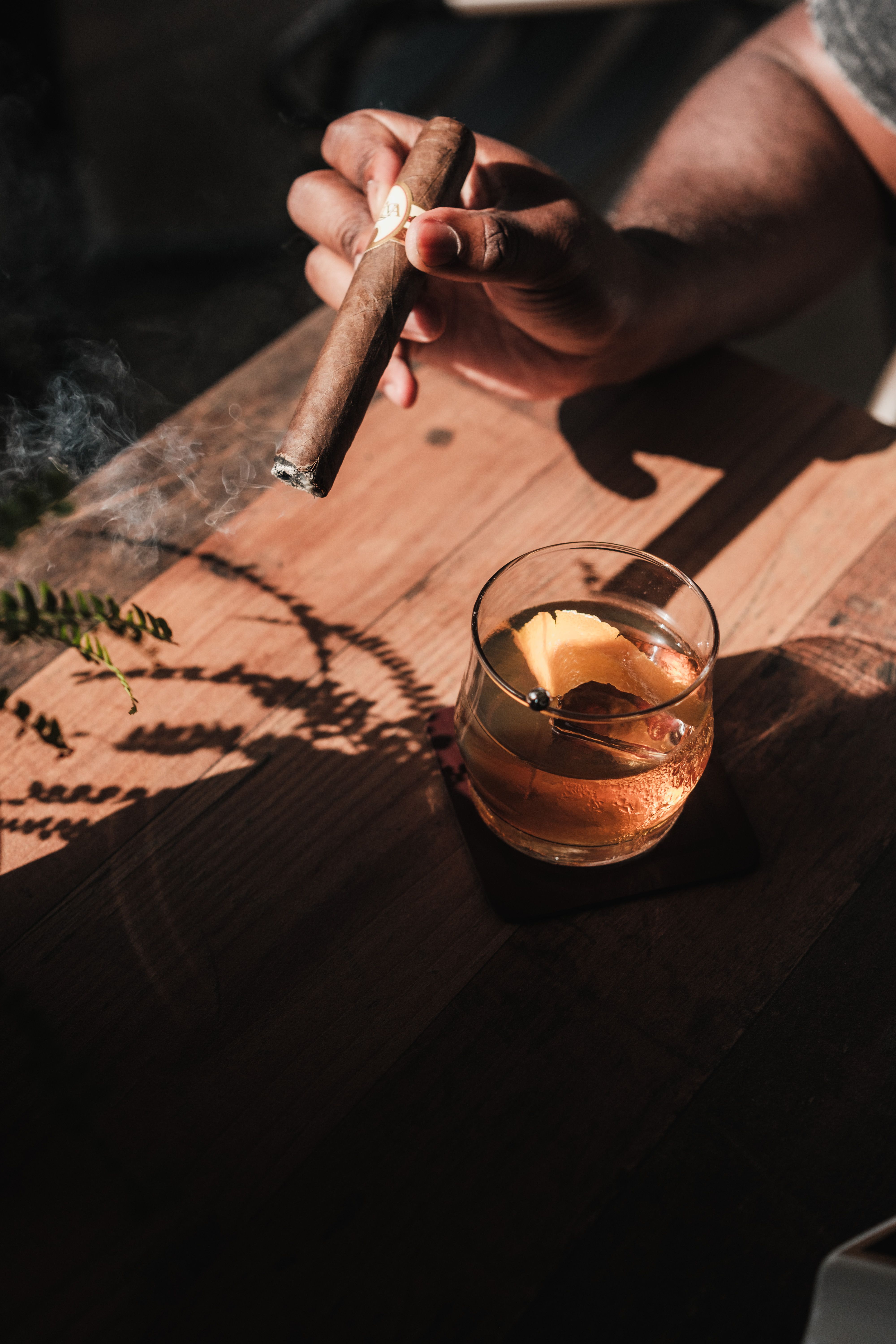 Atmospheric close-up of a hand holding a cigar alongside a classic cocktail in natural light.