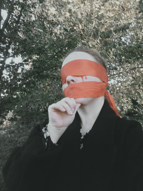 Free A person in a black coat stands outdoors, blindfolded with red tape, surrounded by lush greenery. Stock Photo