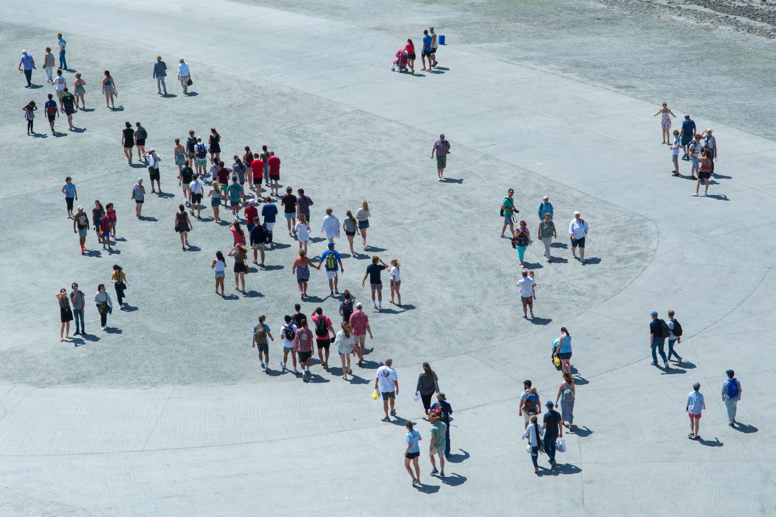 Aerial view of a grey plaza where most people are packed into a dense crowd in the middle, with a scattering of individuals walking alone in the surrounding open space, long shadows stretching behind them