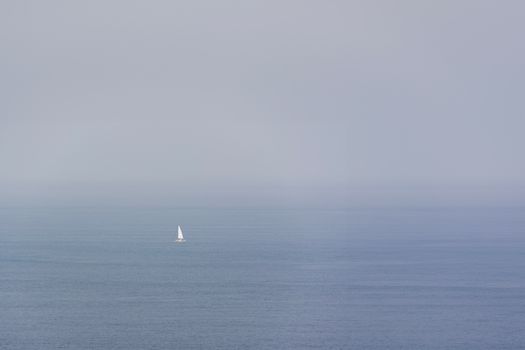 A lone sailboat navigates through a vast, misty ocean under a gray sky.