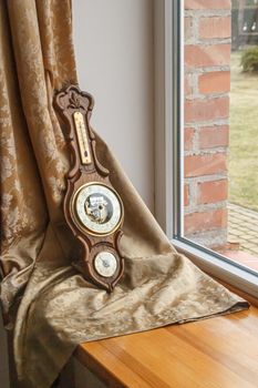Ornate vintage barometer rests elegantly on a wooden window sill with a curtain backdrop.