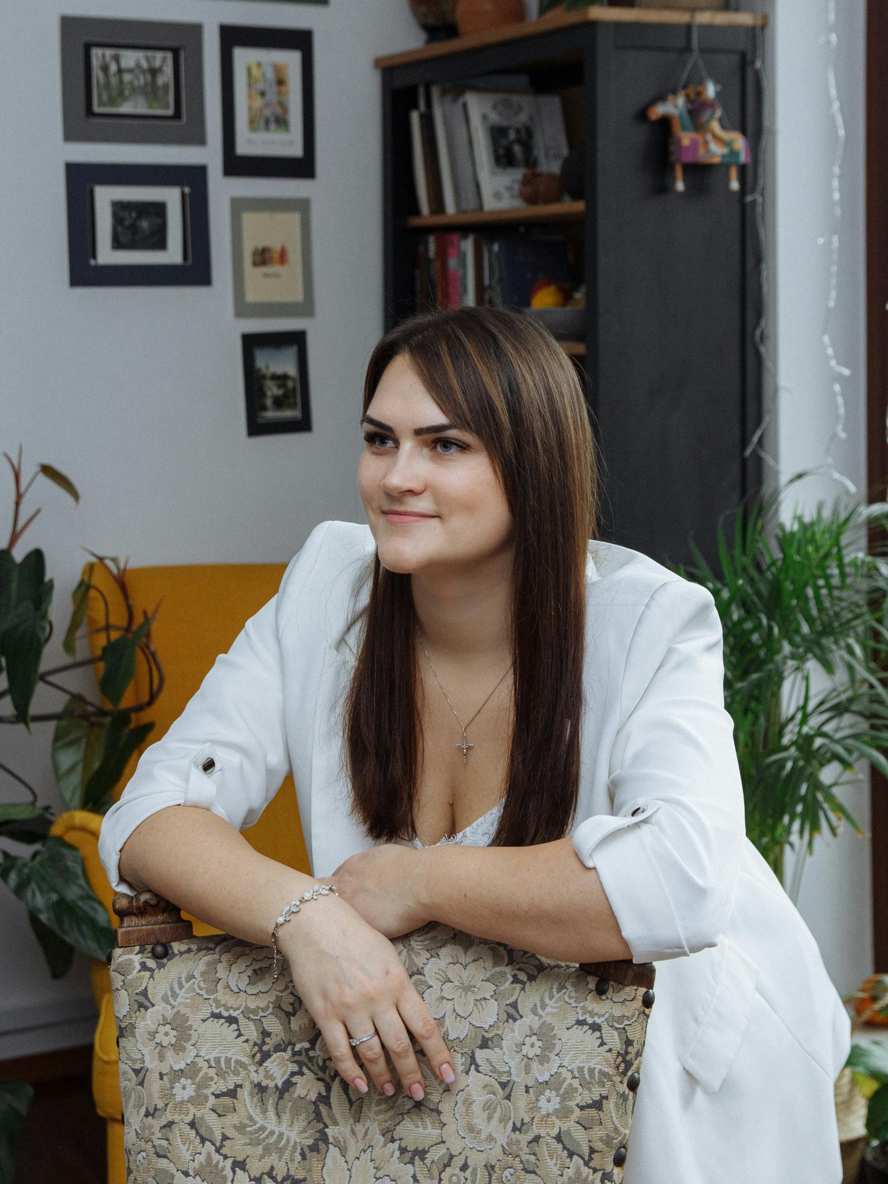 Elegant woman wearing a white suit jacket, sitting casually indoors with a warm smile.