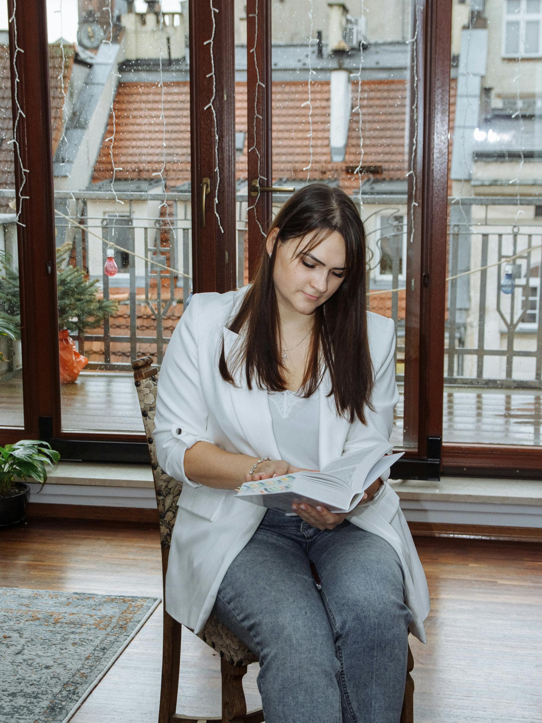Young woman reading a book indoors by a large window with a cityscape view, in casual attire.