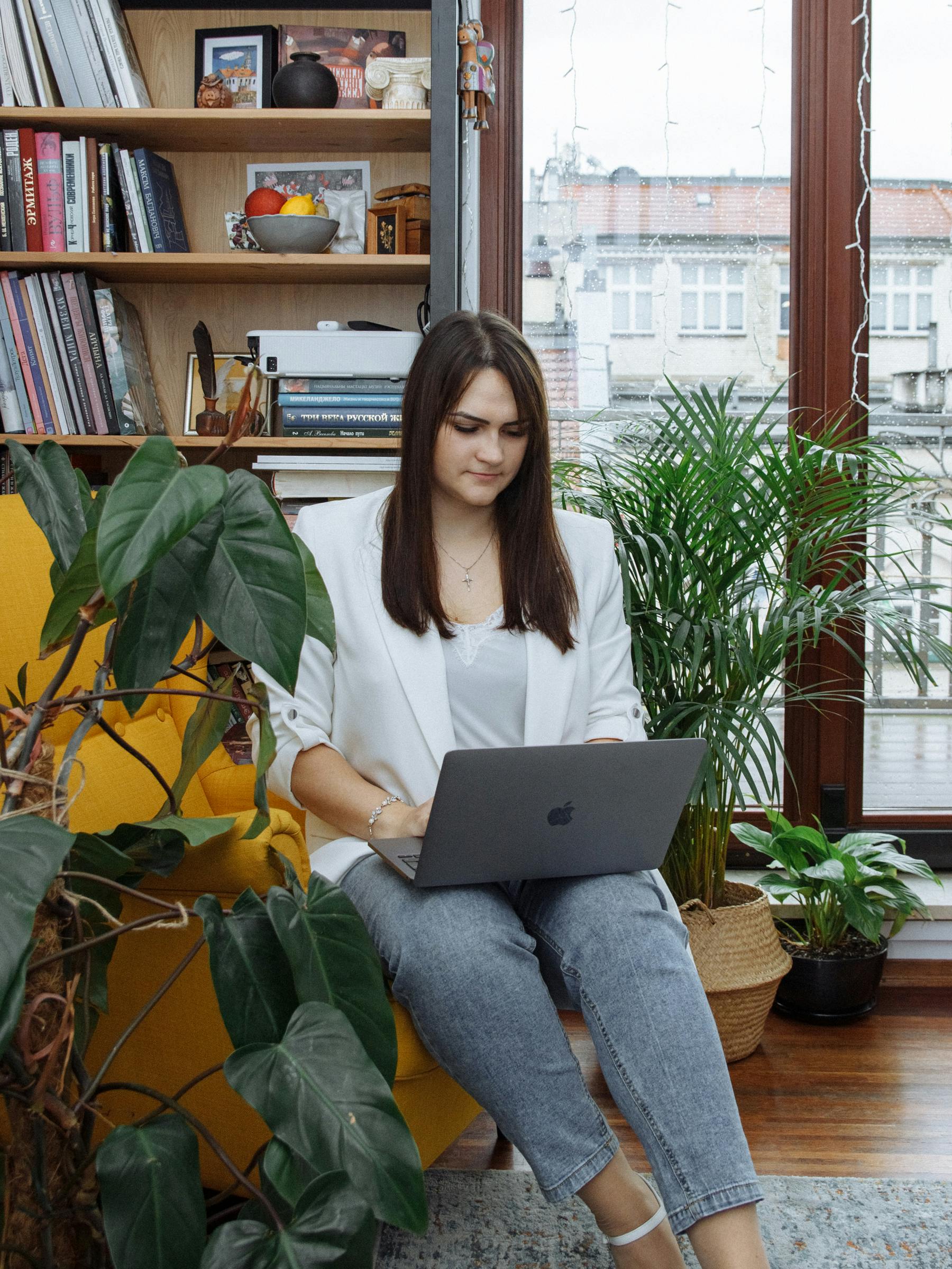 A young woman sits by a window, working on a laptop surrounded by books and plants.