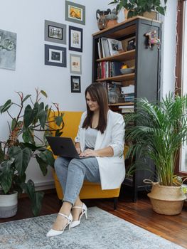A woman using a laptop in a modern home office with plants and elegant decor, showcasing fashion and productivity.