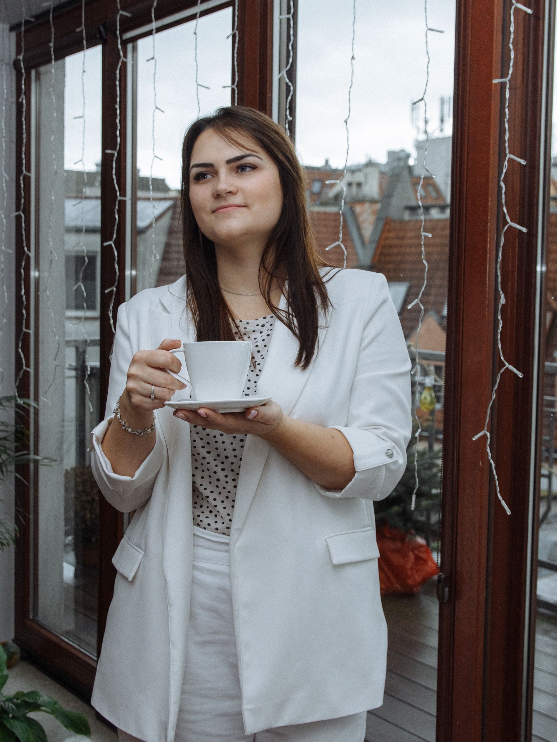 A confident businesswoman in a white blazer enjoys a coffee break indoors by large windows.