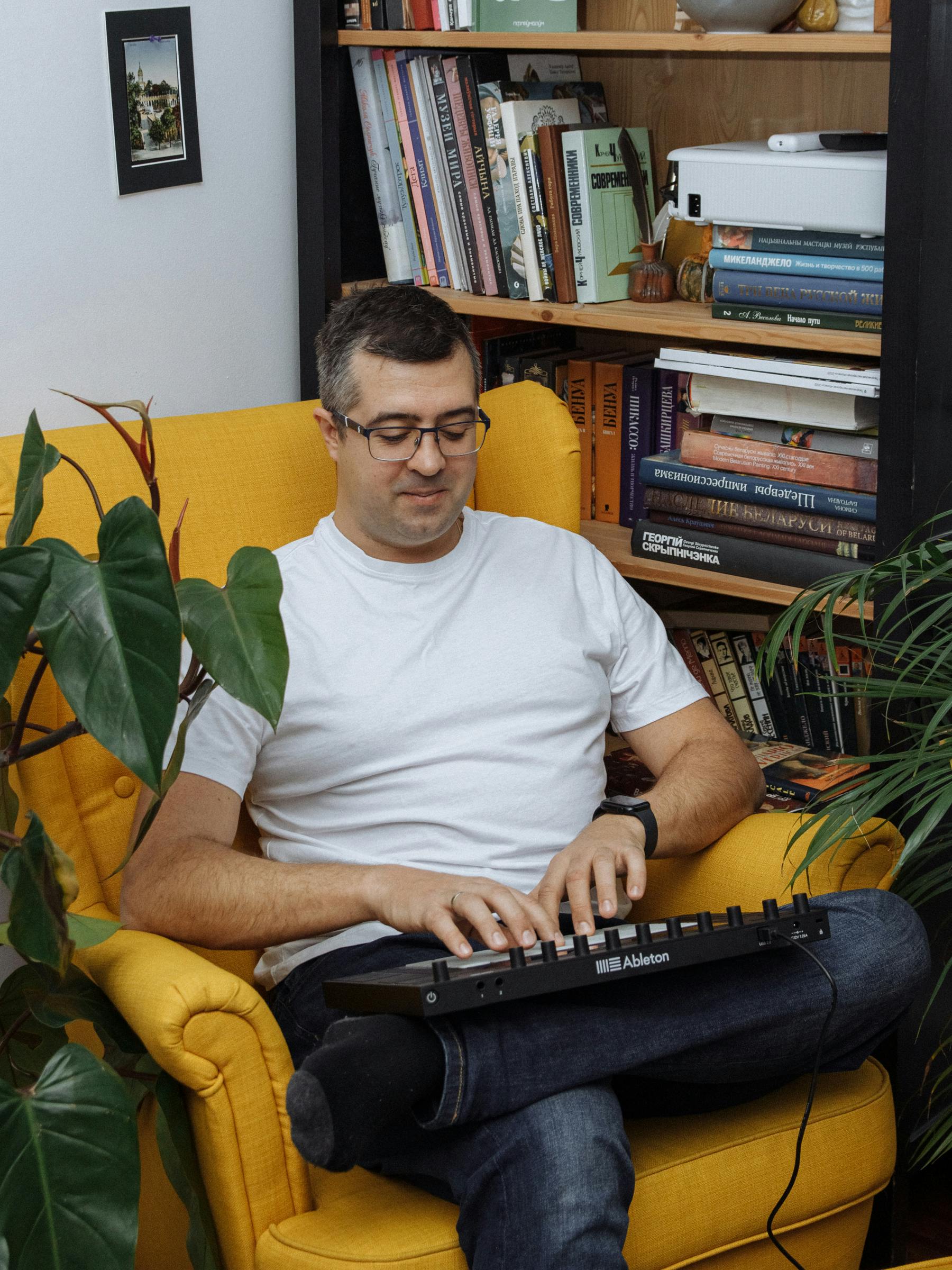 Man sitting on a yellow armchair using a musical keyboard with a bookshelf in the background.
