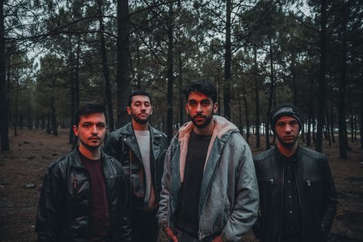 Four men in stylish jackets stand in a tranquil forest setting, exuding a moody vibe.