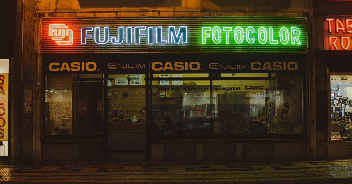 Vibrant neon signs illuminate a Fujifilm and Casio store in Lisbon's night scene.