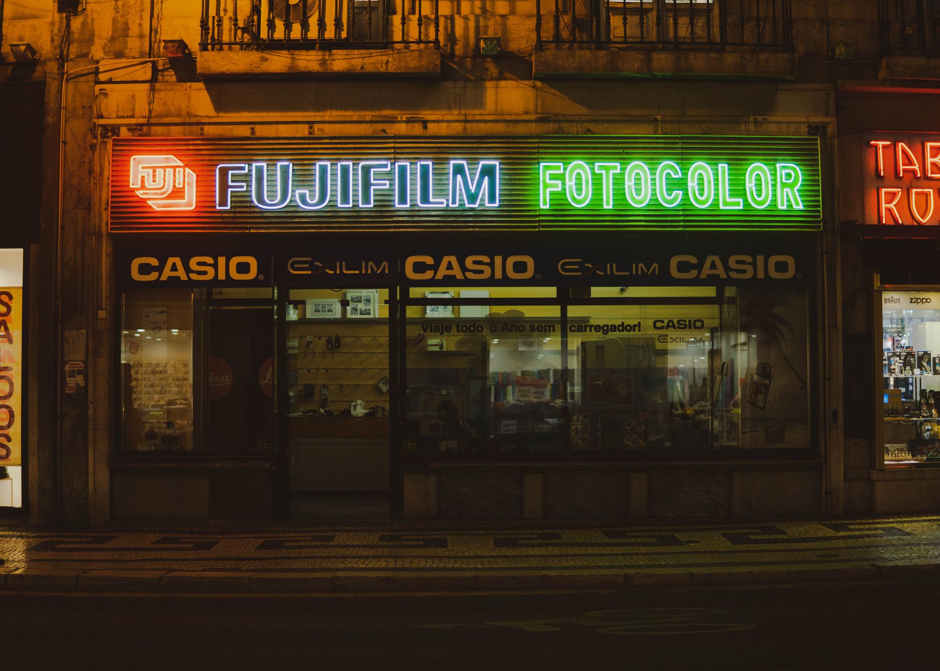 Vibrant neon signs illuminate a Fujifilm and Casio store in Lisbon's night scene.