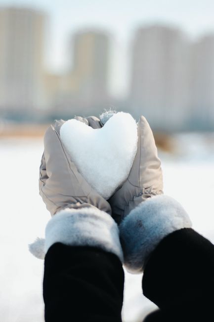 Hands in gloves holding a heart-shaped snowball against city backdrop.