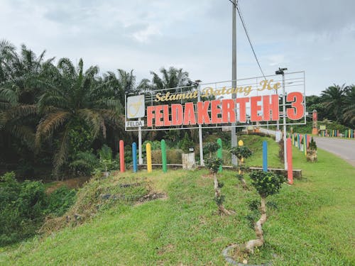 A sign with the text 'Welcome to Felda Kerteh 3' under a cloudy sky near an oil palm plantation.