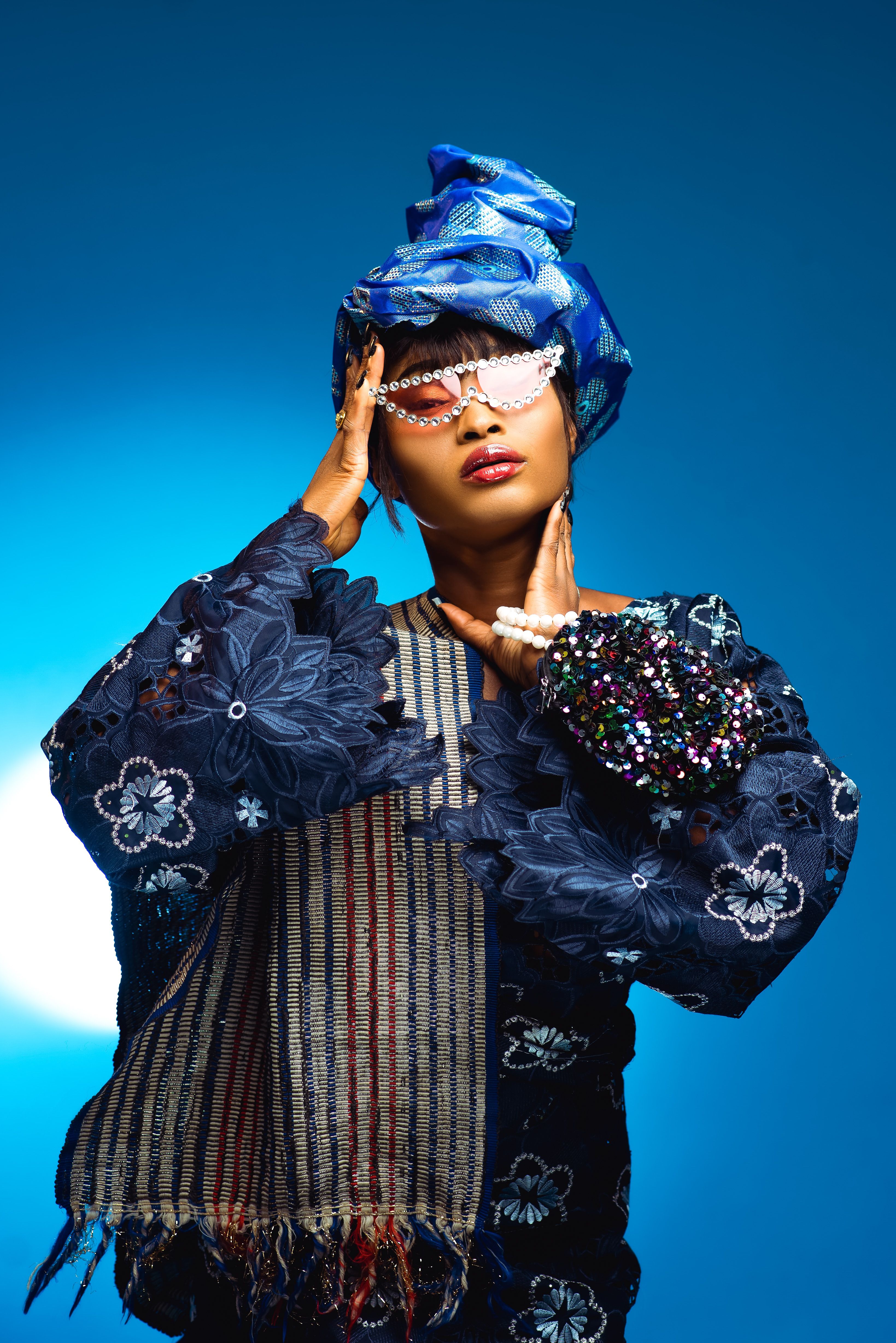 Elegant African woman poses in blue traditional attire with pearl sunglasses, exuding confidence.