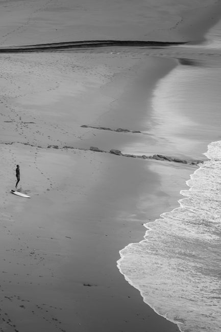 A lone surfer standing on the sandy beach with surfboard, gazing towards the sea in black and white.