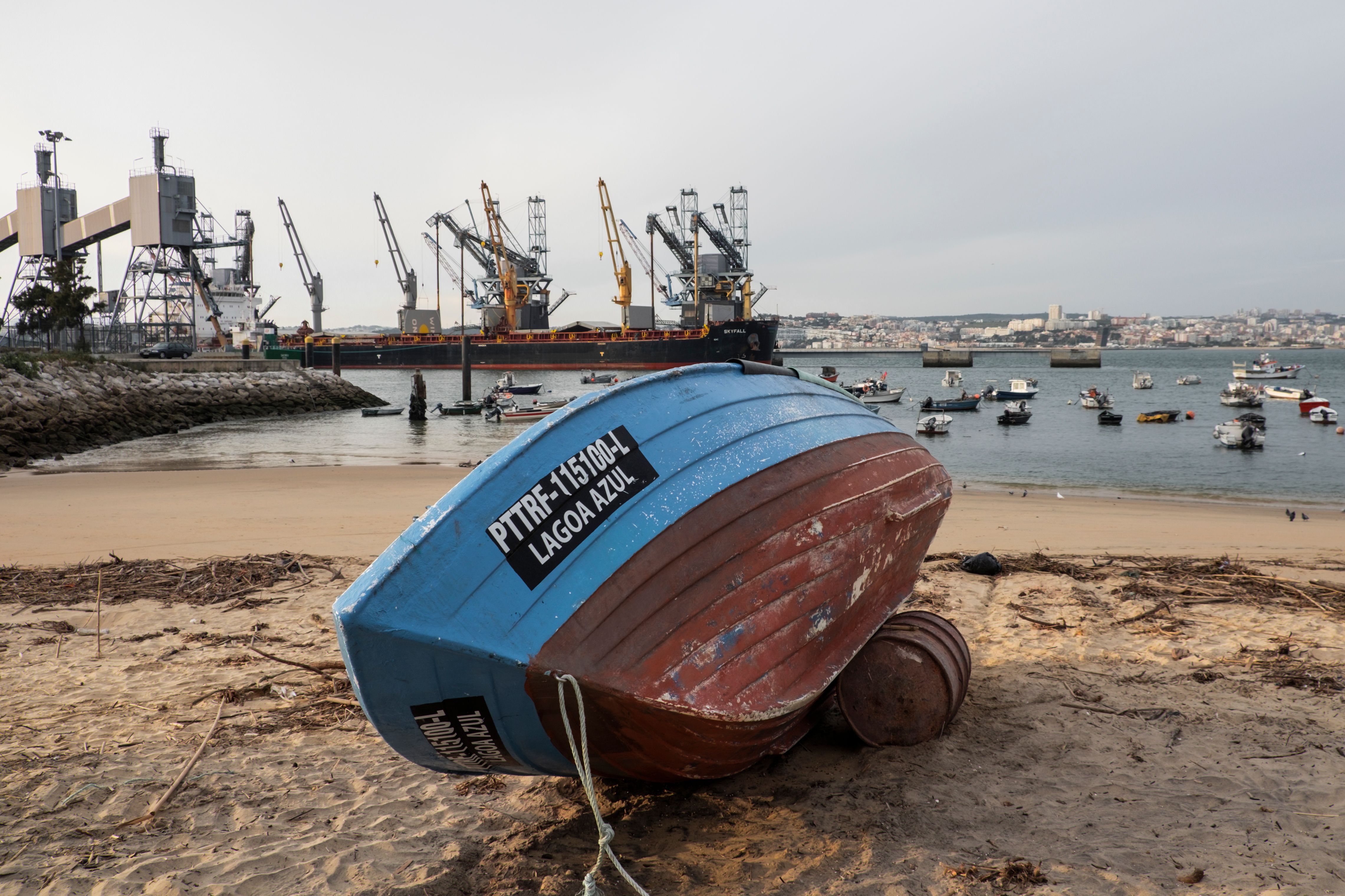 A vibrant boat rests on Trafaria beach with harbor cranes and boats in Setúbal, Portugal.
