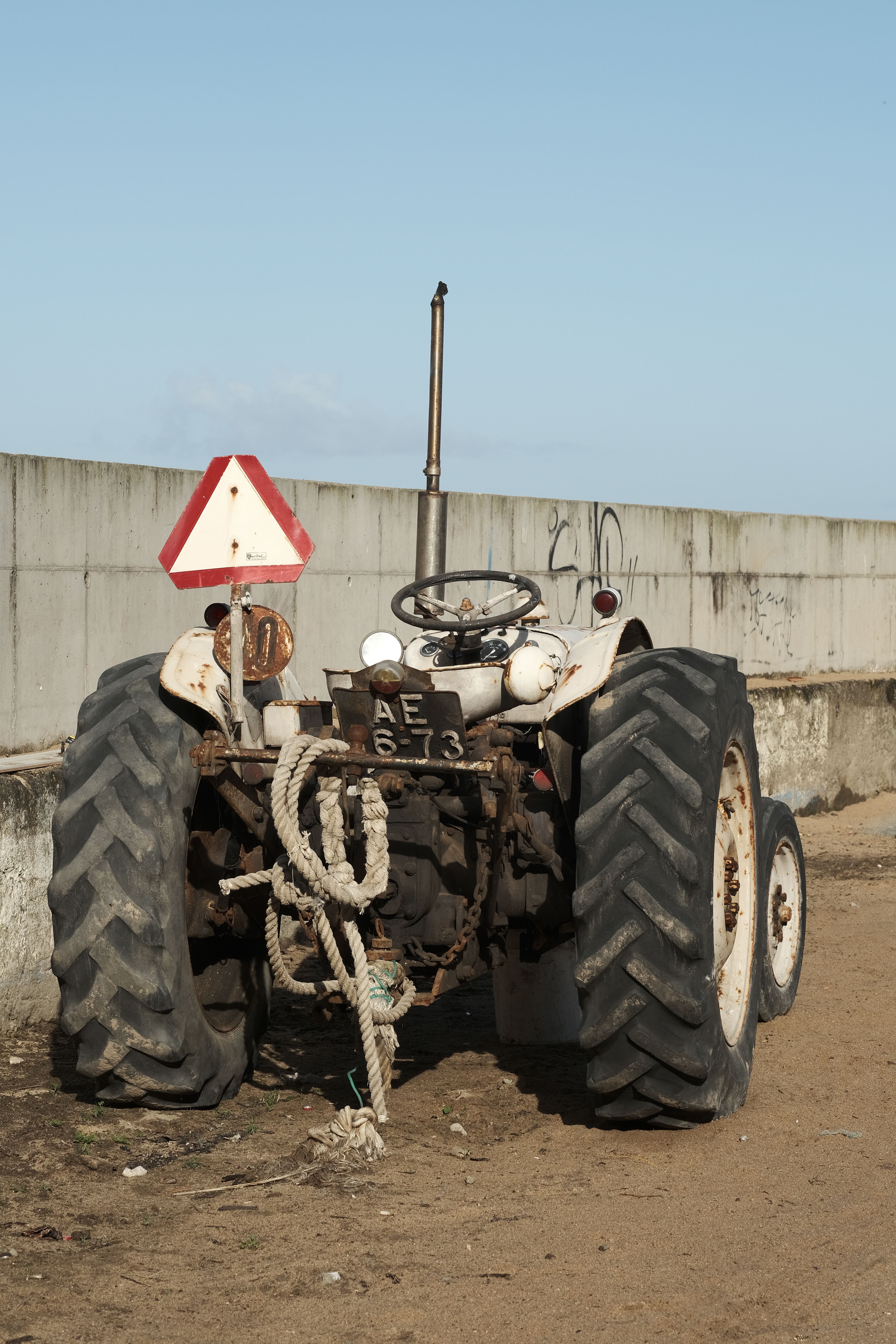 A rusty tractor parked on a dirt road by a concrete wall under clear skies in Ericeira, Portugal.