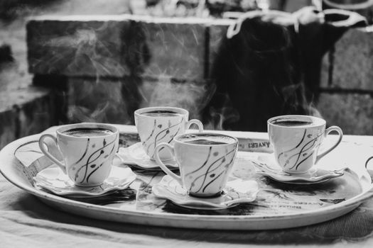 Black and white image of hot coffee cups on a tray, emitting steam, in an outdoor setting.