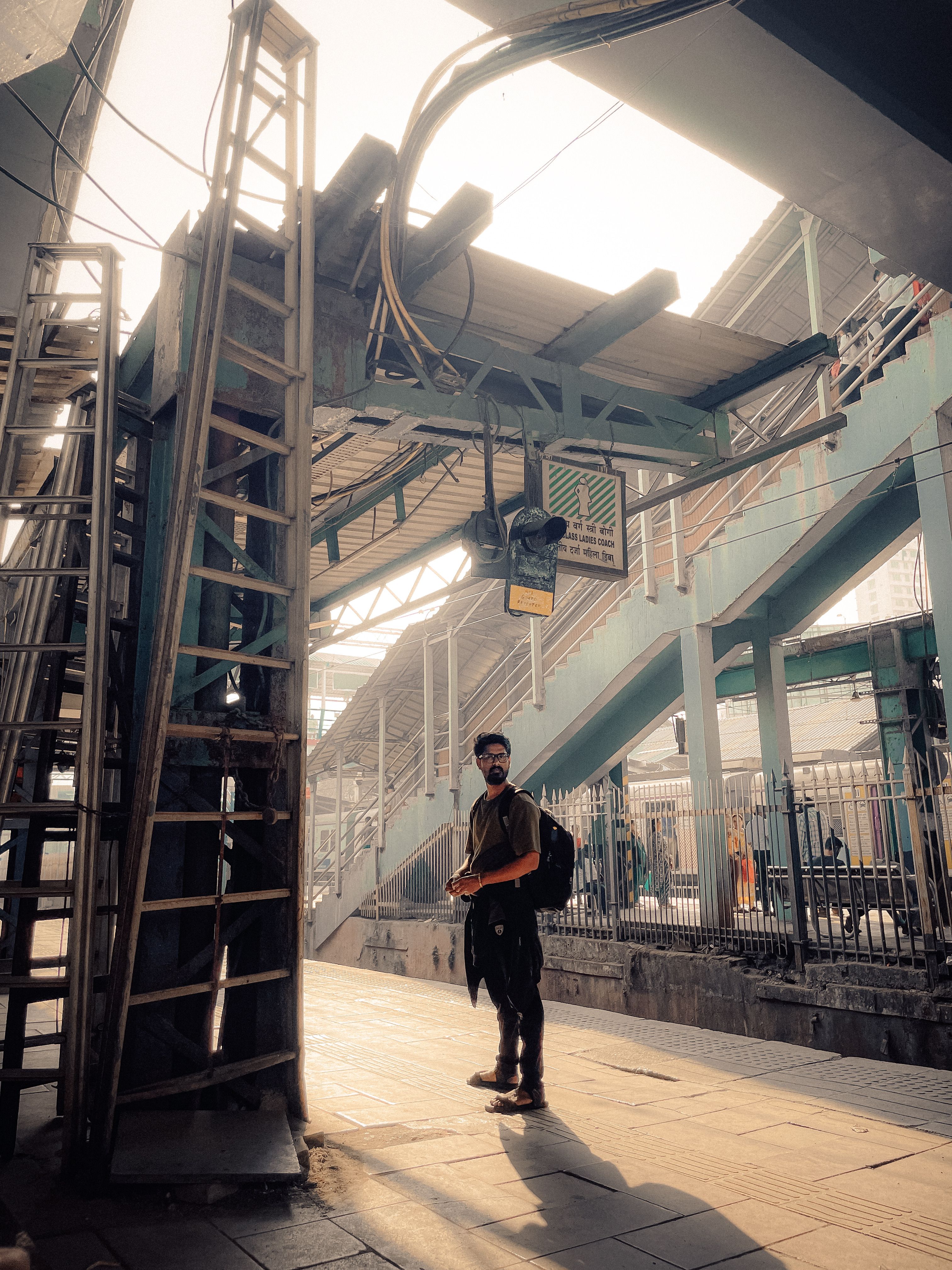 Man with a backpack standing under an overpass at a bustling Mumbai railway station.